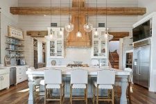 an elegant white barn kitchen with planked walls, stained wooden beams, shaker style cabinets, a vintage kitchen island and white stools