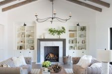 an exquisite barn living room with stained wooden beams, a brick fireplace and built-in bookcases, grey sofas, a wooden coffee table and neutral poufs