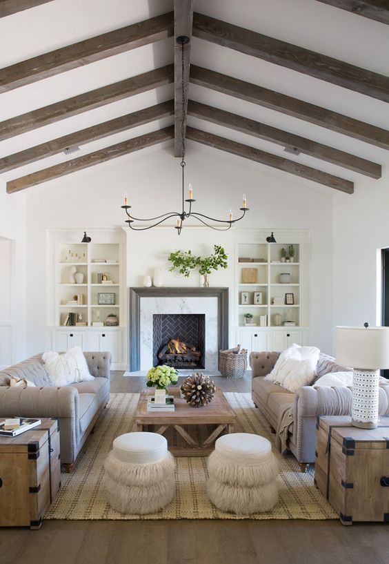 an exquisite barn living room with stained wooden beams, a brick fireplace and built-in bookcases, grey sofas, a wooden coffee table and neutral poufs