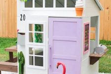 an ice cream inspired kids’ playhouse with a lilac door, a colorful pompom garland, a cactus and a stained dining set behind it