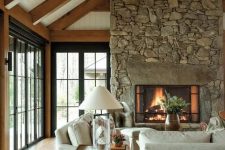 an inviting barn living room with wooden beams, a fireplace clad with stone, neutral seating furniture, wooden tables and some greenery