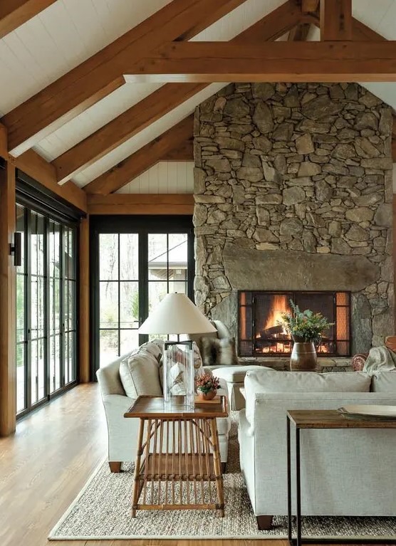 an inviting barn living room with wooden beams, a fireplace clad with stone, neutral seating furniture, wooden tables and some greenery