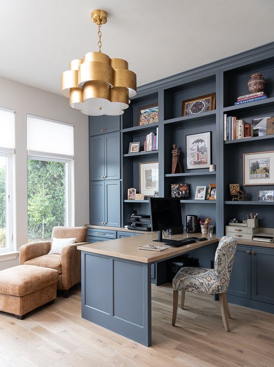 a blue home office with shaker cabinets, a stone countertop on the built in desk, a leather chair with a footrest and a chic gilded chandelier