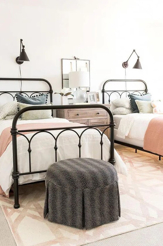 a farmhouse guest bedroom with two metal beds and touches of various muted colors, a polka dot pouf and black sconces over each bed