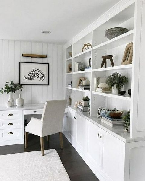 a white farmhouse kitchen with shaker scabinets, open storage units, a built-in desk and a creamy chair plus greenery