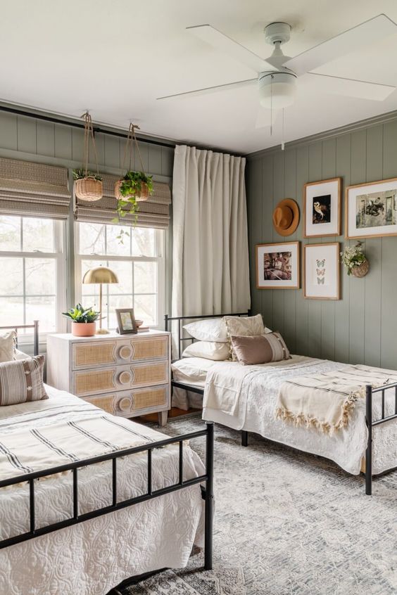 a lovely boho shared guest bedroom with olive green planked walls, black metal beds with neutral bedding, a neutral dresser with cane decor and a gallery wall