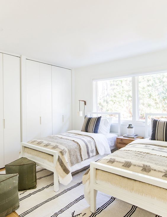 a mid century modern neutral shared guest bedroom with a couple of wardrobes, white beds with neutral bedding,a  striped rug and green stools