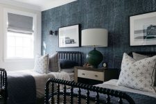 32 a shared guest bedroom with a denim accent wall, black beds with blue and grey bedding, tan stools and a large table lamp