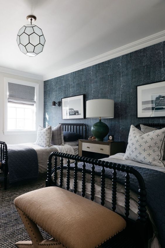 a shared guest bedroom with a denim accent wall, black beds with blue and grey bedding, tan stools and a large table lamp