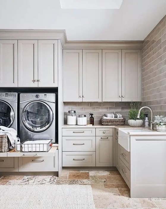 a gorgeous dove grey laundry room done with shaker style kitchen cabinets, a brick wall, potted plants and a cool tile floor