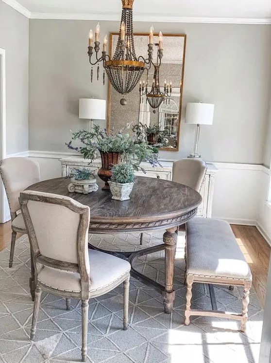 a French country dining room with greige walls, paneling, a fireplace, a stained round table, neutral chairs and a vintage chandelier