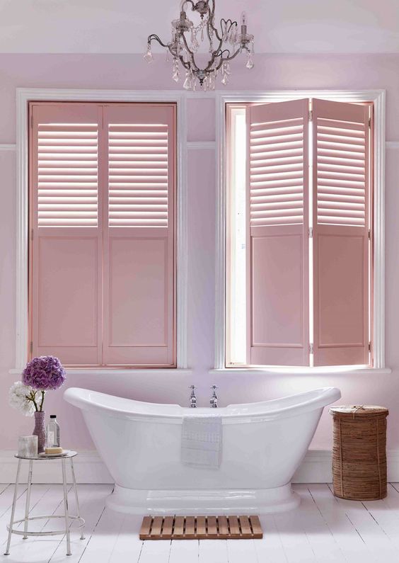 a bathroom with a pink accent wall and pink shutters that highlight the feminine feel, a crystal chandelier and an oval tub
