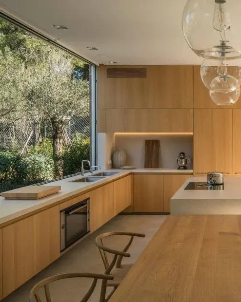 a beautiful contemporary kitchen with sleek light stained cabinets, white stone countertops, built in lights and a glazed wall with garden views