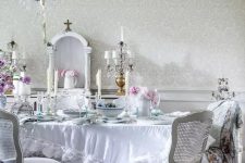 a beautiful vintage dining room with a crystal chandelier, white furniture, a ruffle tablecloth and blooms