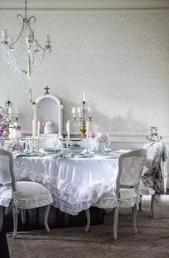 a beautiful vintage dining room with a crystal chandelier, white furniture, a ruffle tablecloth and blooms