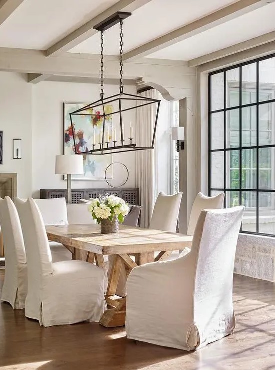a beautiful vintage farmhouse dining room with neutral walls and greige wooden beams, a wooden dining table, upholstered chairs and a chandelier