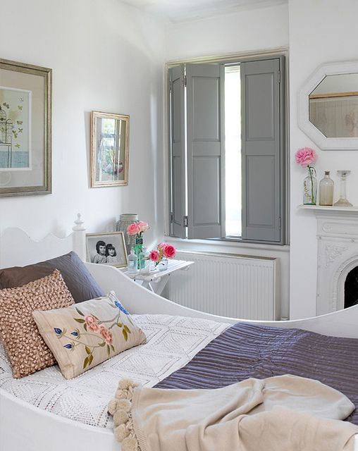 a beautiful white bedroom with touches of vintage, a non working fireplace, a white bed with pastel and white bedding, a white nightstand and some art