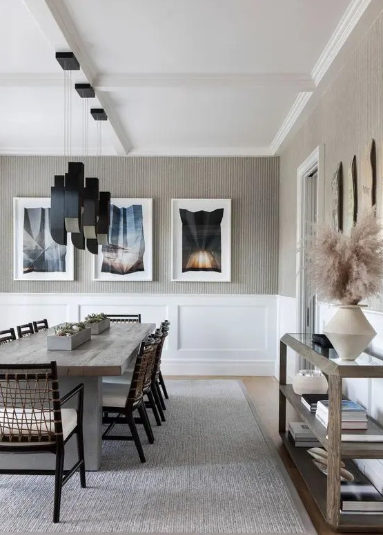 a bold and whimsy greige dining room with white paneling, a wooden table, woven chairs, black pendant lamps, a glass console table and gallery walls