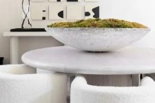 a bold contemporary dining room with a neutral console and a catchy artwork, a round stone table, creamy rounded chairs and a bowl with moss