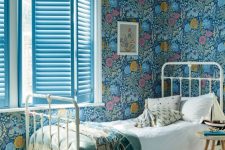 a bold kid’s room with teal floral wallpaper, a white metal bed with neutral bedding, a bold rug and blue shutters
