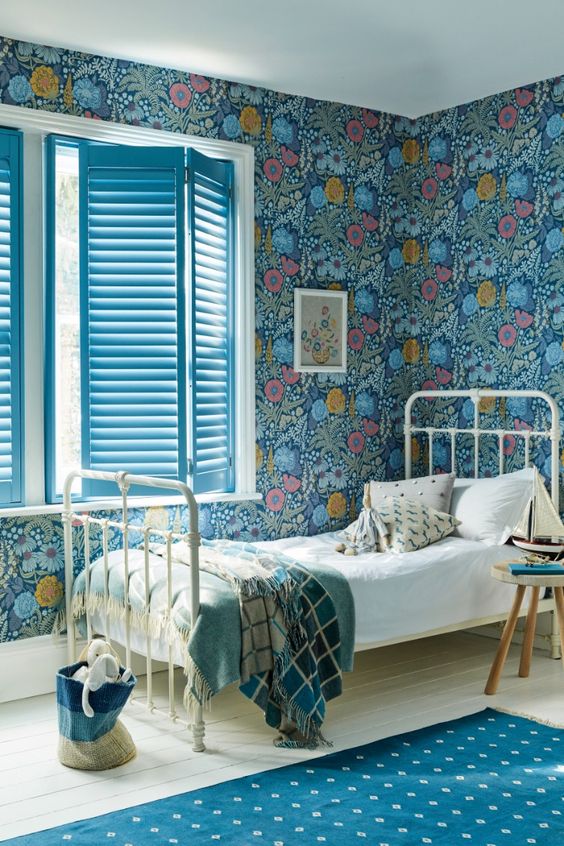 a bold kid's room with teal floral wallpaper, a white metal bed with neutral bedding, a bold rug and blue shutters