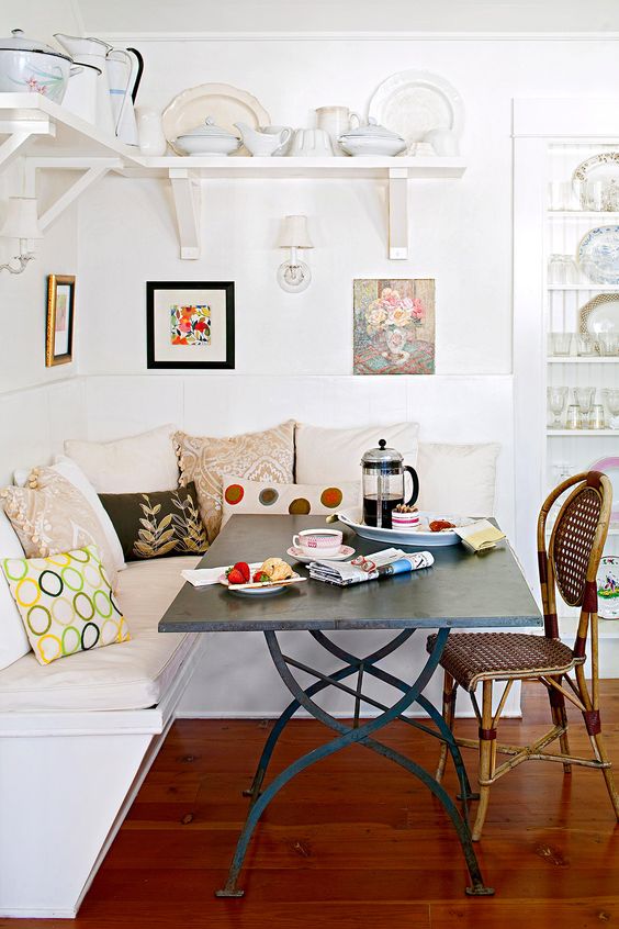 a bright eclectic dining room with a corner bench, a metal table, a rattan chair, some floral artwork and vintage porcelain over the space