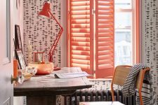a catchy working space with printed wallpaper, a stained vintage desk, an orange plywood chair, a bold red table lamp and coral shutters
