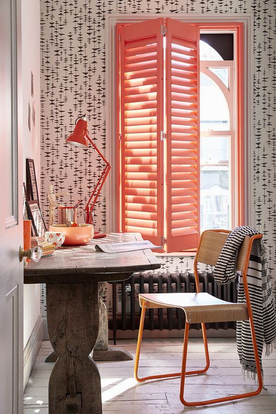 a catchy working space with printed wallpaper, a stained vintage desk, an orange plywood chair, a bold red table lamp and coral shutters