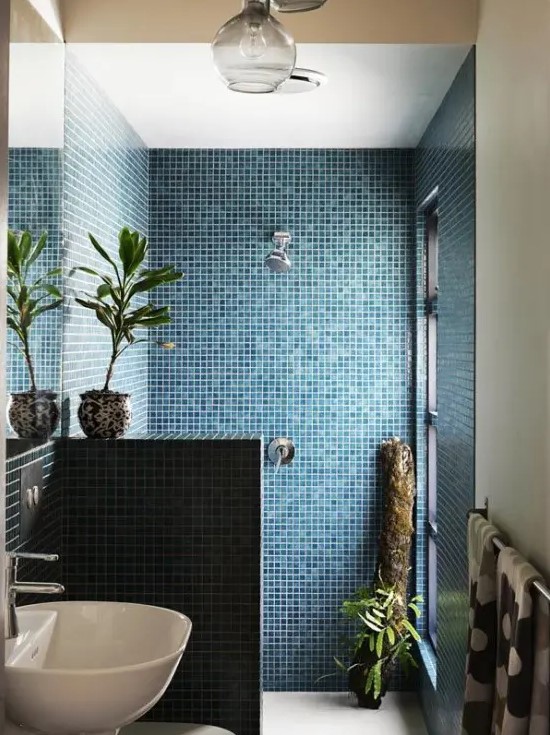 a chic small bathroom clad with blue tiles, with a pony wall and a window in the shower space, potted plants is small and comfy