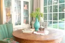 a chic vintage dining space with a glazed wall, a large vintage buffet, a stained round table, a mint green upholstered chair and a floral stool plus a chic chandelier
