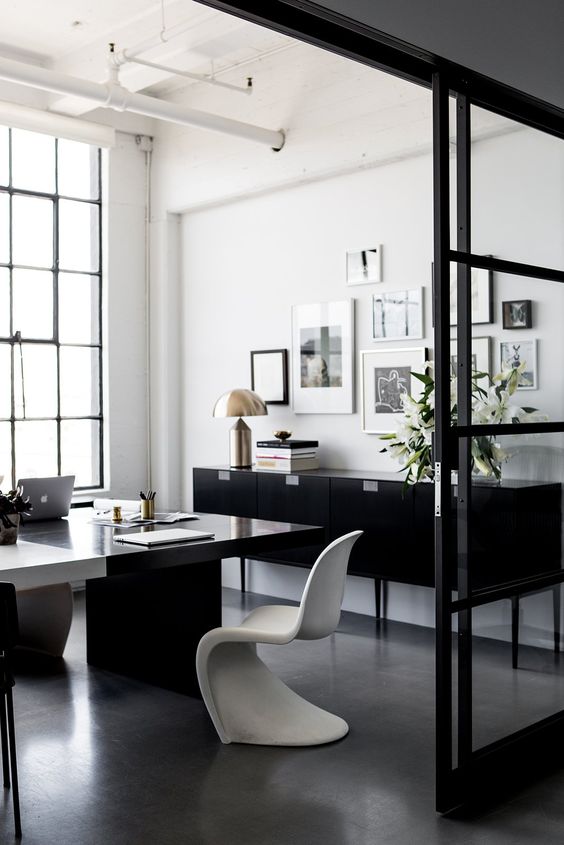 a contemporary black and white home office with a large black desk, a sculptural white chair, a black sideboard, a gallery wall and a table lamp