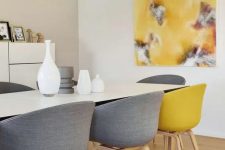 a contemporary dining room with neutral walls, a storage unit, a white table, grey and yellow chairs and a matching artwork