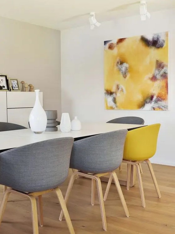 a contemporary dining room with neutral walls, a storage unit, a white table, grey and yellow chairs and a matching artwork