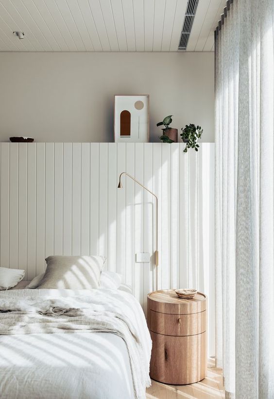 a contemporary light filled bedroom done in creamy shades, with a paneled storage unit, a round wooden nightstand, a brass sonce