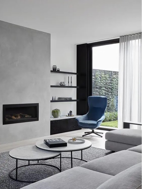 a contemporary living room with a concrete fireplace, built in shelves, a blue chair, a grey sectional and a duo of tables
