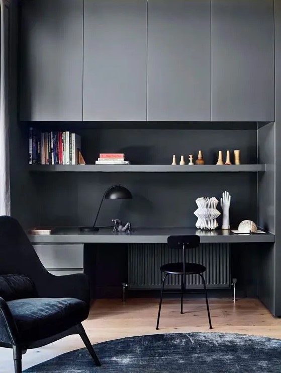 a contemporary moody home office with graphite grey sleek cabinets, an open shelf and a built in desk, a black chair and a navy one, a black table lamp