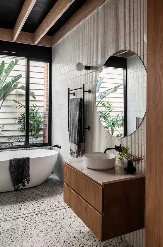 a contemporary tropical bathroom with a white terrazzo floor, an accent wall with skinny tiles, a floating vanity, a round sink and a tub