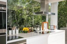 a contemporary white kitchen with sleek cabinetry and stone countertops plus glazed walls that open on the garden