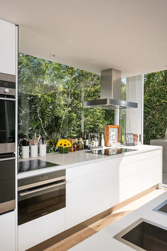 a contemporary white kitchen with sleek cabinetry and stone countertops plus glazed walls that open on the garden