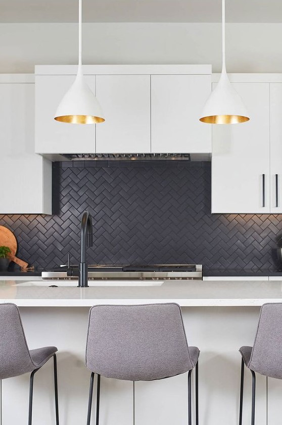 a contemporary white kitchen with a black herringbone backsplash, tall grey stools and pendant lamps with gold inner