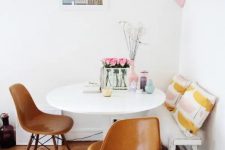 a cool and chic dining area with a bench with printed pillows, a white round table, leather chairs, some blooms and pastel accessories