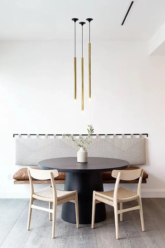 a cool modern dining area with a floating bench with a soft back, a black round table, woven chairs and a lovely gold tube lamp