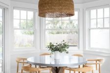 a cool neutral modern boho dining room with a round table, woven chairs, a large woven pendant lamp and greenery in the vase