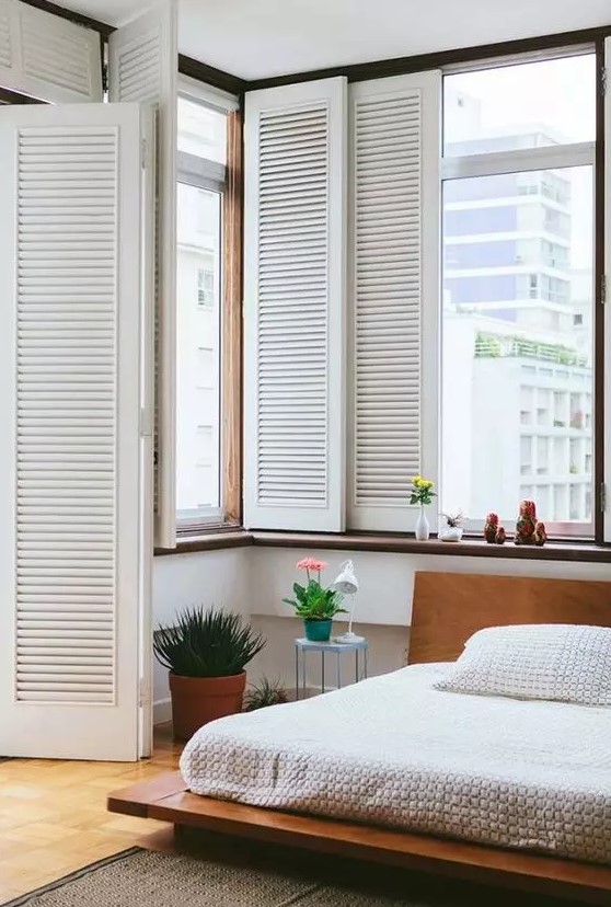 a cozy mid century modern bedroom with a low stained bed with neutral bedding, some potted plants and blooms, shutters on the windows