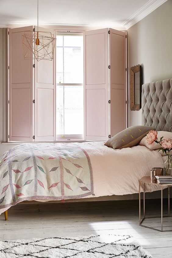 a delicate and welcoming bedroom with grey walls, a grey upholstered bed, blush and grey bedding, pink solid shutters and a catchy pendant lamp