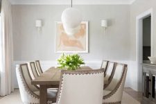 a delicate greige farmhouse living room with greige walls, white paneling, a wooden dining table, creamy upholstered chairs and a dark stained credenza