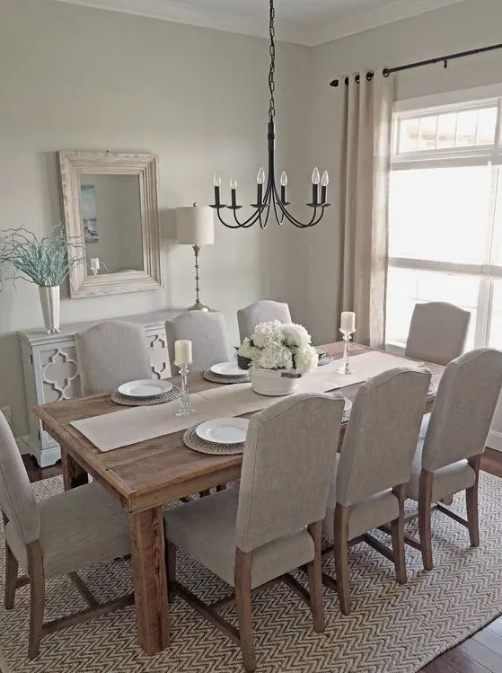 a dreamy greige dining room with a wooden dining table, neutral chairs, a black chandelier, a mirror in a whitewashed frame and a neutral sideboard