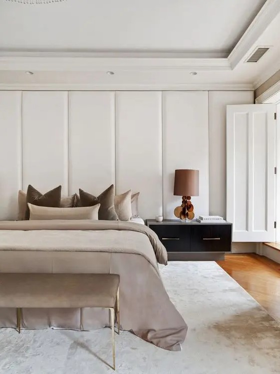 a fabulous contemporary bedroom with a headboard clad with upholstered panels, a bed with neutral earthy bedding and a matching bench, a large nightstand with a lamp