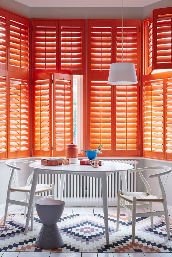 a gorgeous and bright dining room with an oval white table and chairs, a printed rug and super bold orange shutters covering the bay window