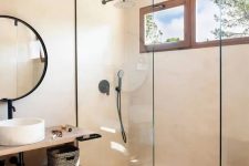 a greige contemporary bathroom with a shower space enclosed in glass, a floating open shelf vanity, a round mirror in a black frame, black fixtures